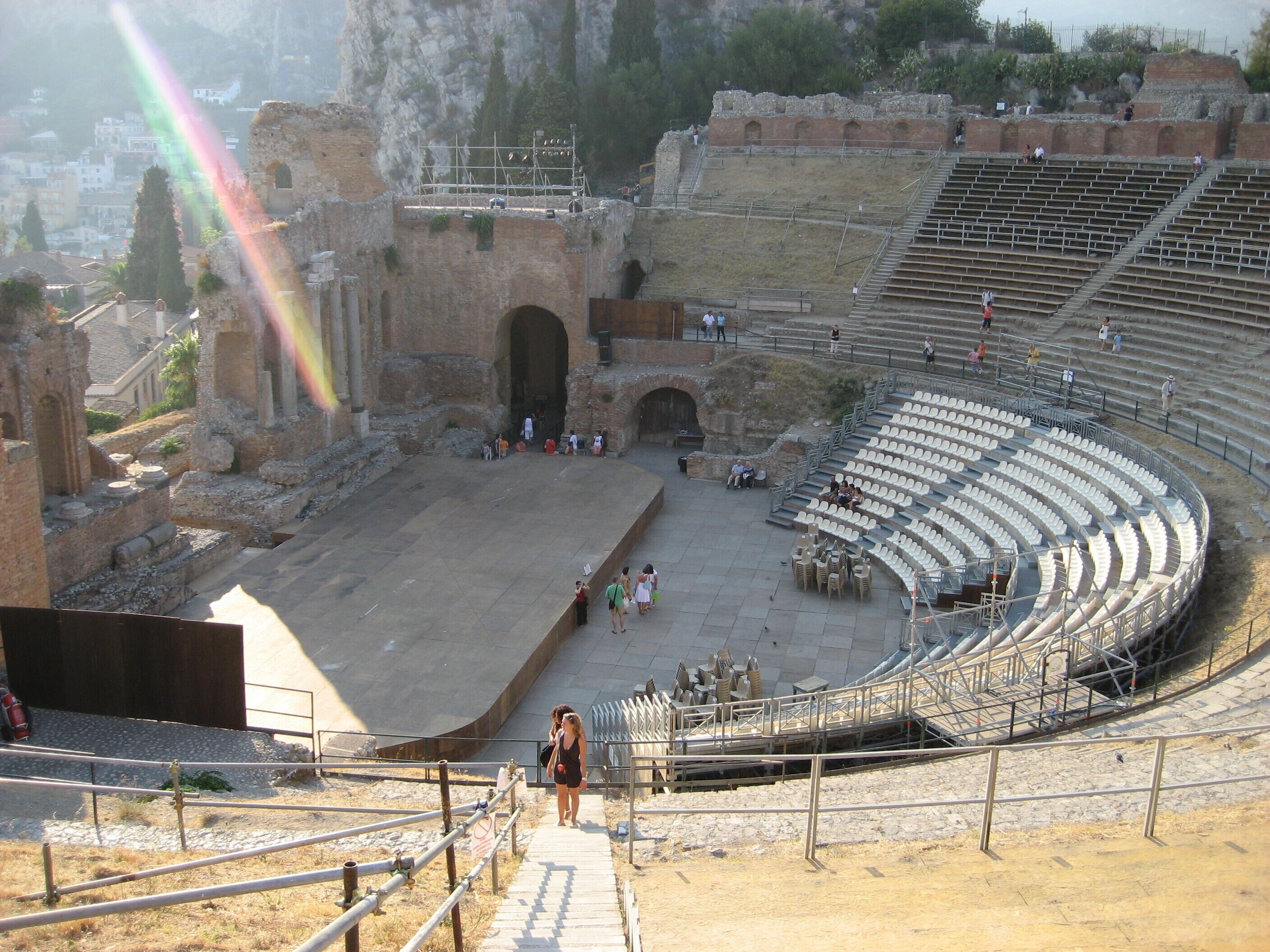 An ancient amphitheater with stone seating and a central stage, surrounded by ruins and modern scaffolding.