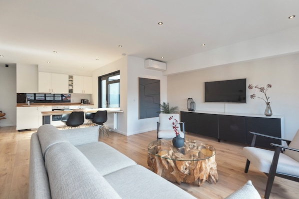 A modern living room with a sofa, a glass coffee table, and a dining area with chairs and a kitchen in the background. - Sack-Time 2: Comfortable stay in a prime location. (Zandvoort)