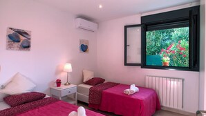 Et hotelværelse med to senge, et vindue med udsigt til grøn natur, en radiator og et lille bord med lampe og blomst.