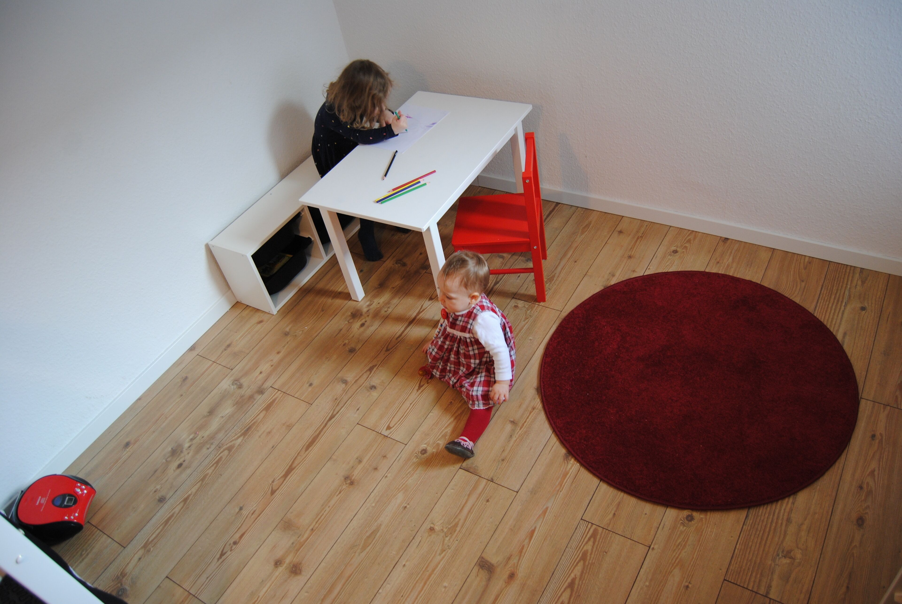 A child sitting at a white desk with a red chair, a red rug, and wooden flooring.