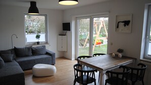 Uma sala de estar moderna com um sofá seccional cinza, uma mesa de jantar de madeira com cadeiras pretas, um banquinho branco e uma luminária pendente preta.