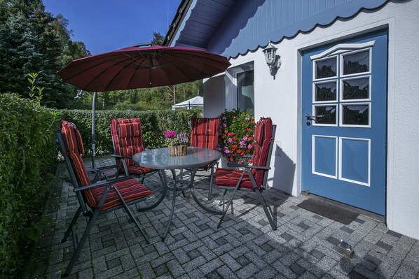 Une terrasse extérieure avec une table ronde, quatre chaises à rayures, un parasol rouge et une porte bleue.