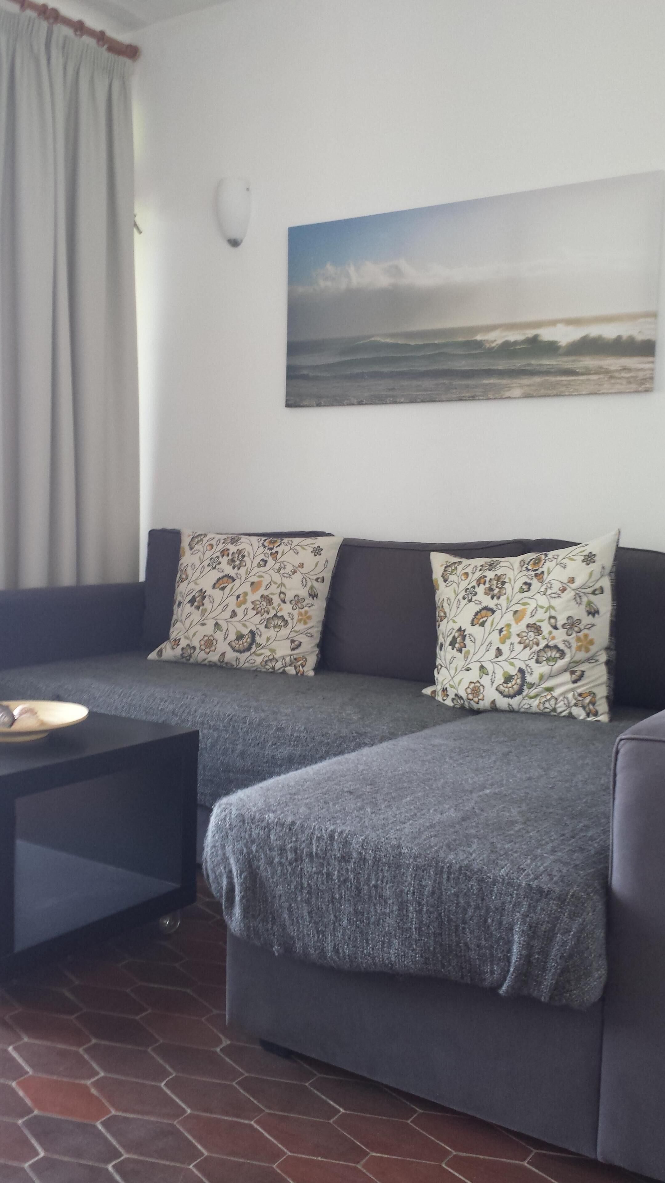 A grey sofa with floral cushions, a small black table, and a painting of a beach scene on the wall.