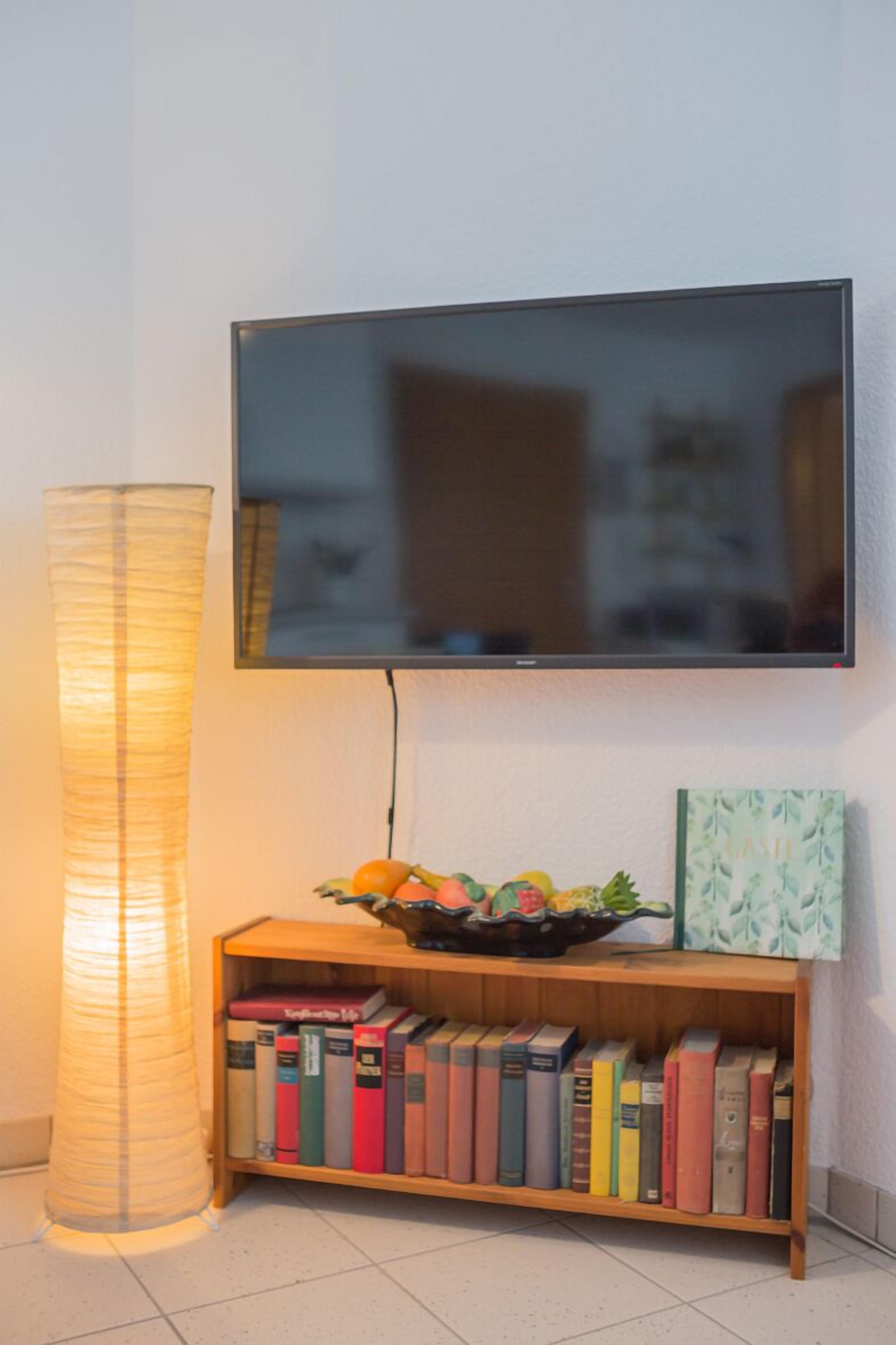 Un téléviseur à écran plat fixé au mur, une bibliothèque en bois remplie de livres, un bol décoratif contenant des fruits et un grand lampadaire cylindrique.