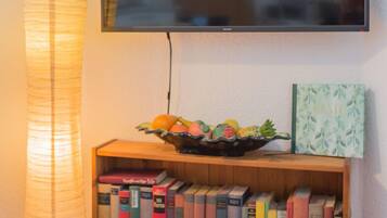 Ein flachbildfernseher an der Wand, ein Bücherregal aus Holz mit Büchern, eine dekorative Schale mit Früchten und eine hohe, zylindrische Stehlampe.