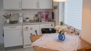 Una cocina con armarios blancos, una mesa de madera con mantel de encaje, un florero azul con flores y una lámpara colgante.