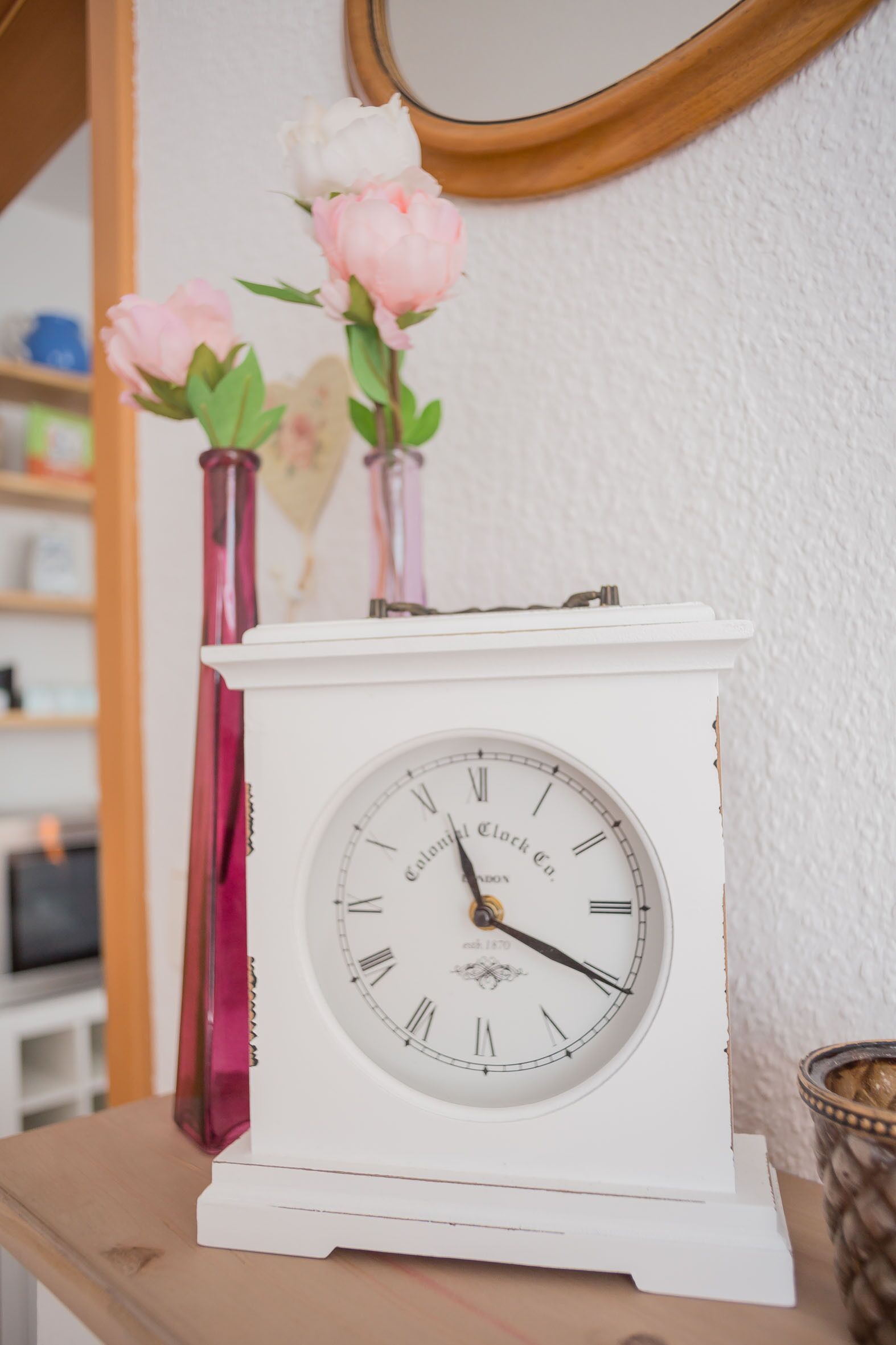 Une horloge blanche avec des chiffres romains et le nom de la marque inscrit sur le cadran, posée sur une surface en bois.