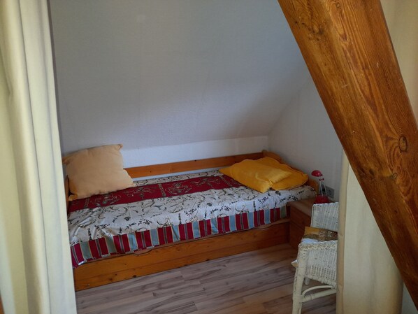 A small, cozy room with a single bed, a wooden headboard, a red and white patterned bedspread, and a yellow pillow.