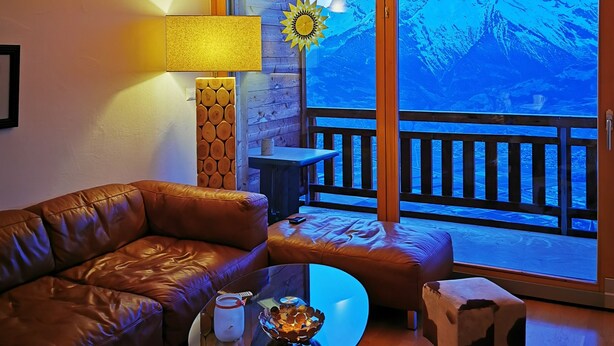 A cozy living room with a leather couch, a coffee table with a lamp, and a large window overlooking a mountain view. - Stylish loft with a great panoramic view (Veysonnaz)