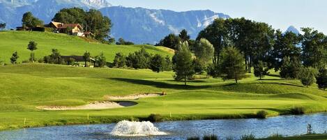 Un campo de golf con un estanque, árboles y montañas al fondo.