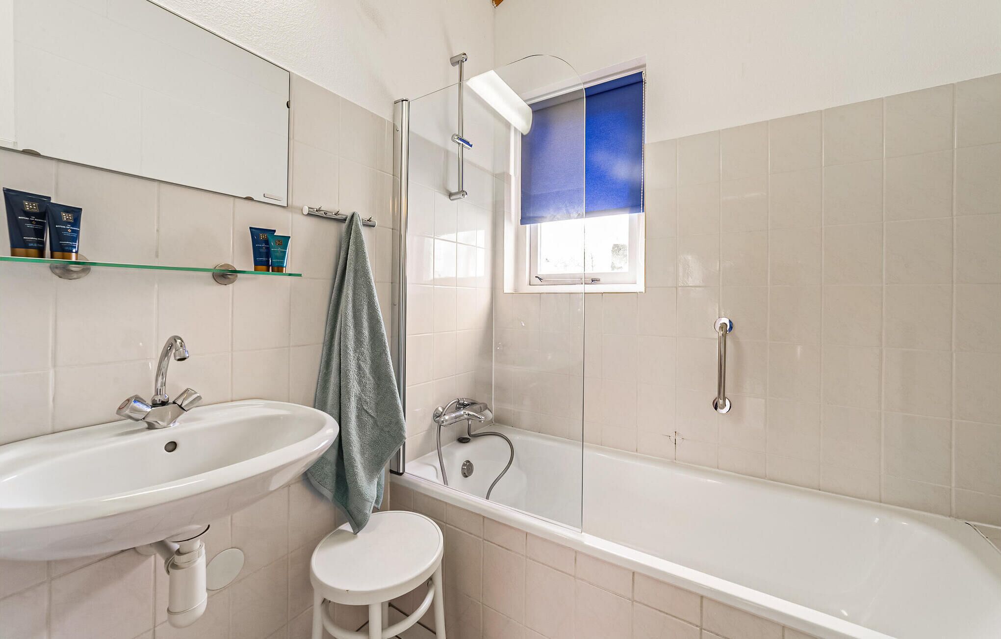 A bathroom with a white sink, a bathtub, and a shower. There is a window with a blue blind, a towel hanging on a rack, and toiletries on a shelf.