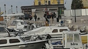 Un puerto deportivo con varias embarcaciones amarradas, un edificio amarillo al fondo y algunas personas caminando.