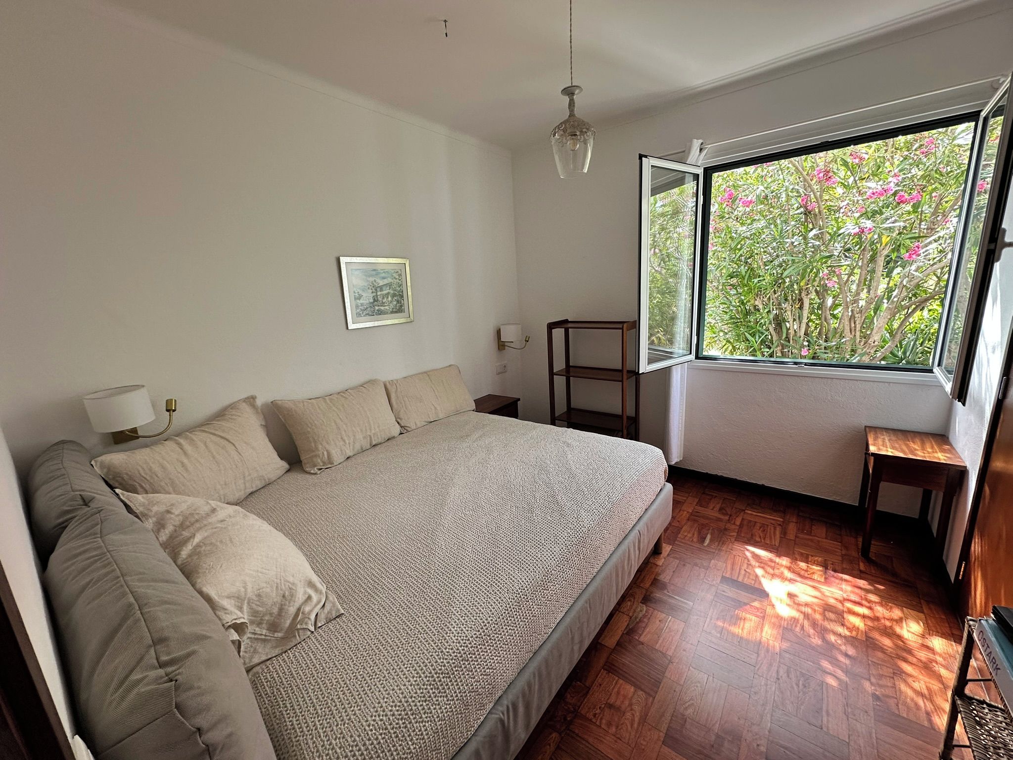 Un dormitorio con una cama grande, una mesita de noche, una lámpara, un cuadro en la pared y una ventana con vistas a zonas verdes.