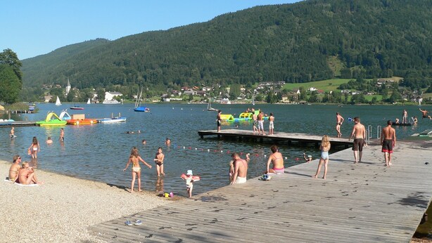 Une scène au bord d’un lac, avec une jetée en bois, des personnes qui se baignent et des bateaux sur l’eau.