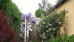 Un chemin de gravier mène à une porte en bois, elle-même entourée d’une végétation luxuriante et de plantes en fleurs.