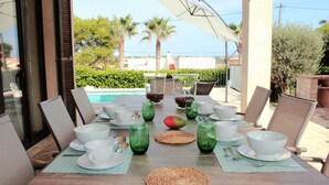 A dining table set for a meal with white plates, bowls, and green glasses, overlooking a pool and palm trees.