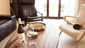 A living room with a wooden coffee table, a bottle of wine, and two glasses. There is a black leather chair and a white chair with a wooden armrest.