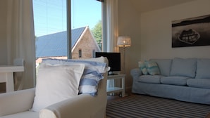 A living room with a white armchair, a blue sofa, a TV on a white stand, and a lamp. - Lakeside new direct lakeside location lakeside terrace, bathing lake, biosphere wifi (salem)