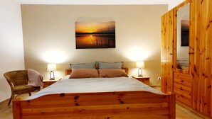 A bedroom with a wooden bed, two bedside tables with lamps, a wicker chair, a large mirror, and a sunset painting on the wall.