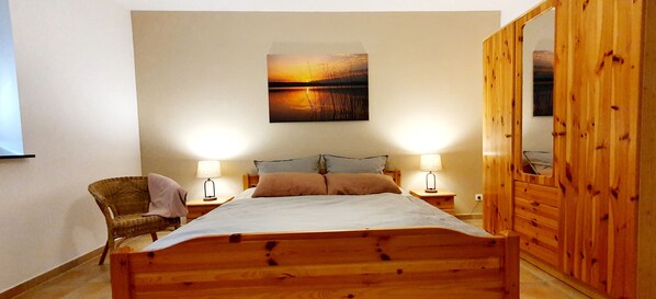A bedroom with a wooden bed, two bedside tables with lamps, a wicker chair, a large mirror, and a sunset painting on the wall.