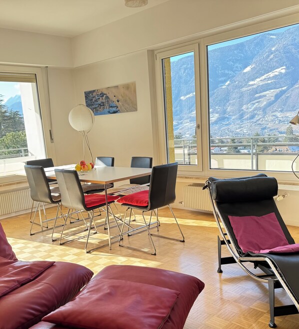 A dining area with a table and chairs, a sofa, and a view of mountains through large windows. - stylish, sunny, and quiet penthouse with a dream view - relax and enjoy (Meran)