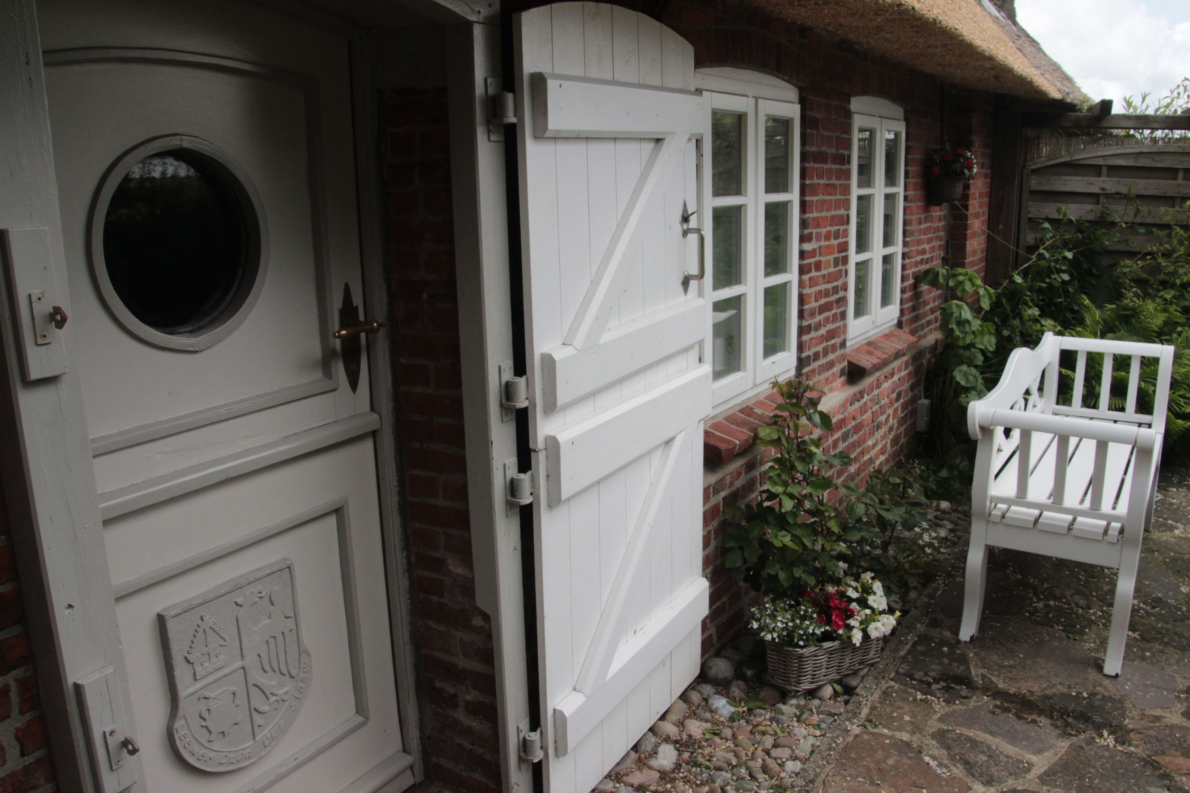 Een witte houten deur met een rond raam en een familiewapen, een wit hek en een witte bank voor een gebouw met rieten dak.