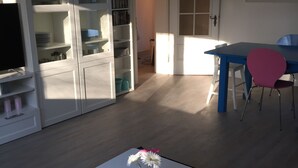 A living room with a white coffee table, a patterned rug, a dining area with a blue table and pink chairs, and a kitchen area with open cabinets and a TV. - NEW! Modern 3-room apartment in Lüneburg, close to the city, quiet, outskirts (Lüneburg)