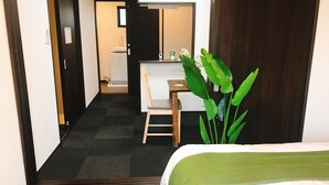 A hotel room with a bed, a desk, and a chair. There is a plant in the corner and a door leading to another room. - GH One More Heart at Kamishichiken 3 (Kyoto)