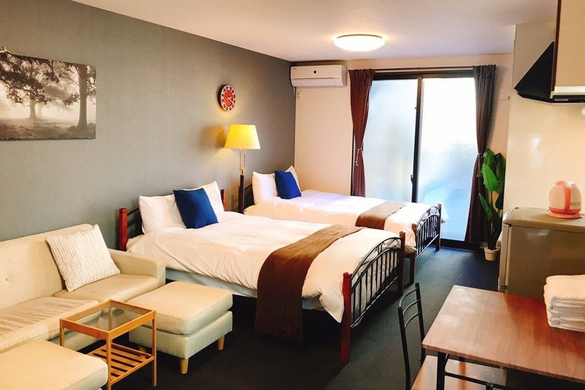 A hotel room with two beds, a sofa, a small table, a lamp, and a window with curtains.