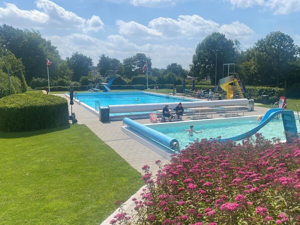 Un espace piscine comprenant plusieurs bassins, un toboggan et une végétation luxuriante tout autour.
