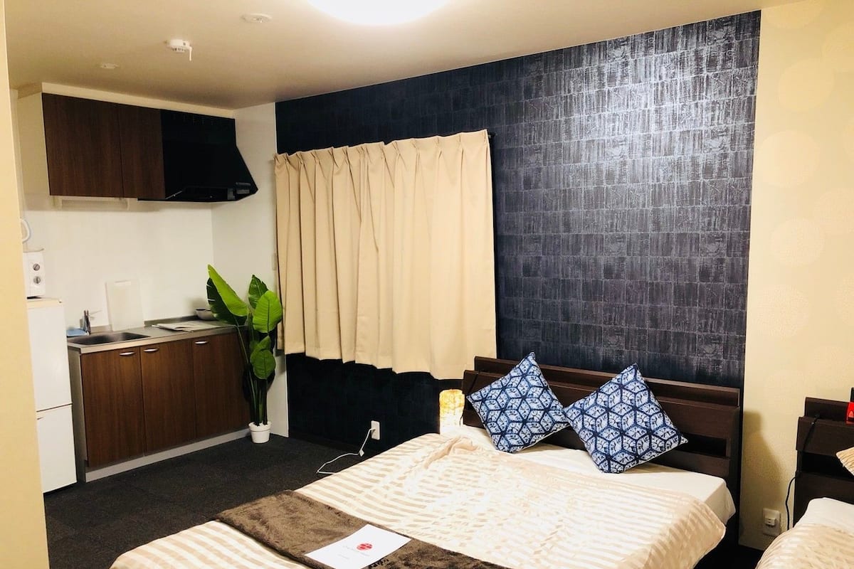 A hotel room with a bed, a small kitchenette, a dark textured wall, and a window with curtains.