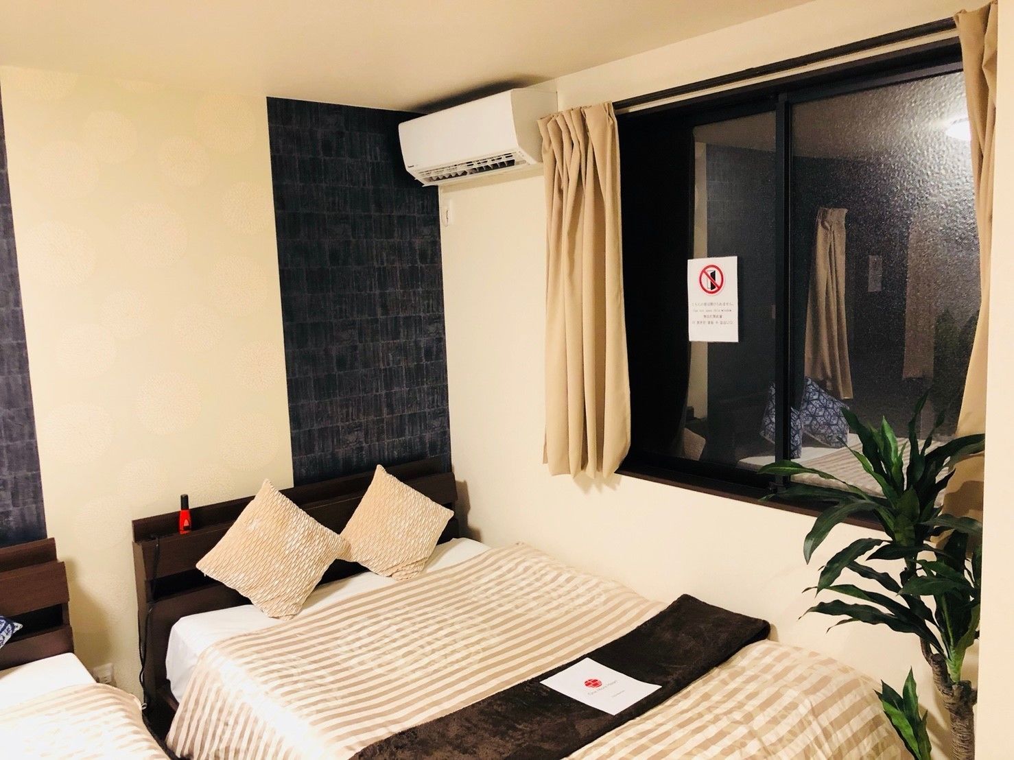 A hotel room with two beds, a dark brick accent wall, an air conditioning unit, a window with curtains, and a plant.
