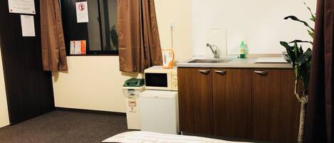 A hotel room with a bed, a small kitchenette, a window with curtains, and a plant.