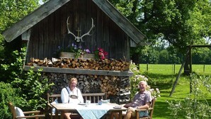 Two people seated at a wooden table with a blue tablecloth, outside a rustic wooden structure with a deer head decoration and stacked firewood. - Heissbauer - Cozy "Obstgarten" Vacation Rental with Mountain Views - Ideal for Families (Wackersberg)