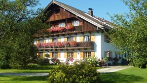 A traditional chalet-style building with multiple balconies adorned with flowers, surrounded by lush greenery. - Heissbauer - Cozy "Obstgarten" Vacation Rental with Mountain Views - Ideal for Families (Wackersberg)