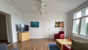 A living room with a wooden floor, a TV on a wooden stand, a red sofa, a blue rug, and a door in the background.