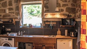 Une cuisine rustique avec un mur en pierre, un lave-linge, un évier et une fenêtre donnant sur un paysage verdoyant.