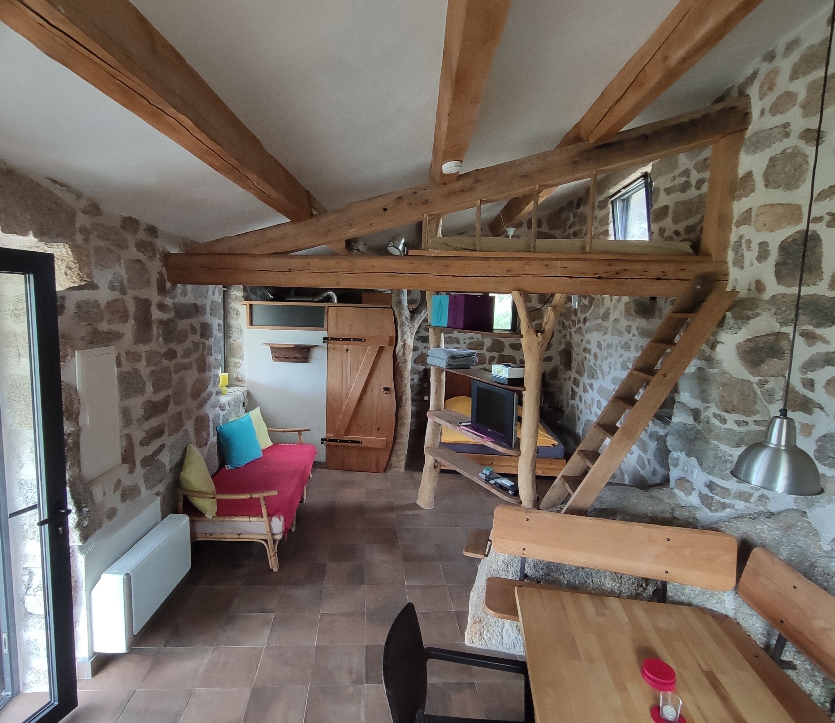 A rustic room with exposed wooden beams, stone walls, a bed with colorful pillows, a small table with a glass, and a ladder.