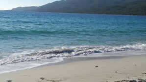 Une plage avec une eau d’un bleu limpide, un rivage sablonneux et des montagnes en arrière-plan.