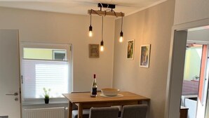 Een eethoek met een houten tafel en vier stoelen, een radiator, een raam met zonwering en een plafondlamp.