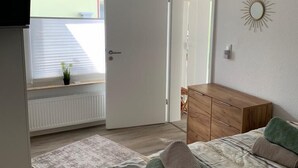 Een slaapkamer met een bed, een houten dressoir, een radiator, een raam met jaloezieën en een plafondlamp.