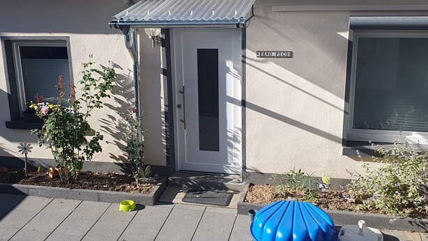 Een woongebouw met een witte deur, twee ramen, een kleine tuin met planten en bloemen, en een blauwe paraplu op het portaal.