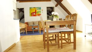 Ein Essbereich mit einem Holztisch und Stühlen, einem Korbstuhl, einem Bild an der Wand und einer Vase mit Blumen.
