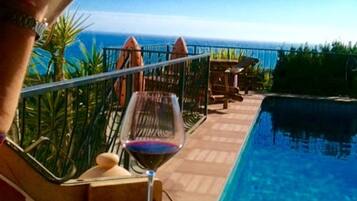 Un área junto a la piscina con una copa de vino tinto, una mesa de madera y vista al océano.