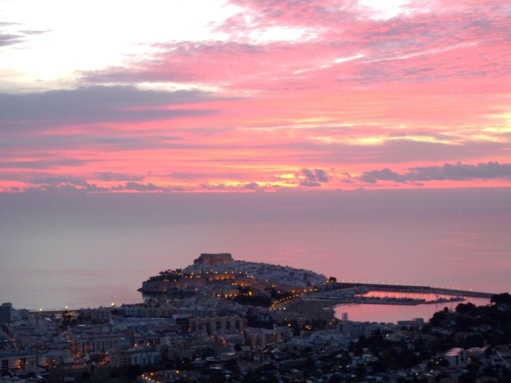 A coastal cityscape at dusk with a prominent hilltop structure, a marina, and a bridge in the distance.