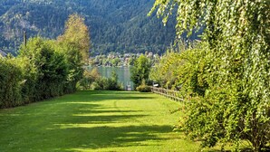 Un sentier herbeux bien entretenu mène à un plan d’eau, entouré d’une végétation luxuriante et offrant une vue lointaine sur un petit village.