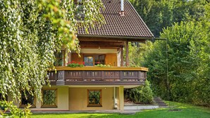 Une maison à deux étages dotée d’un balcon, entourée d’une végétation luxuriante et d’arbres.