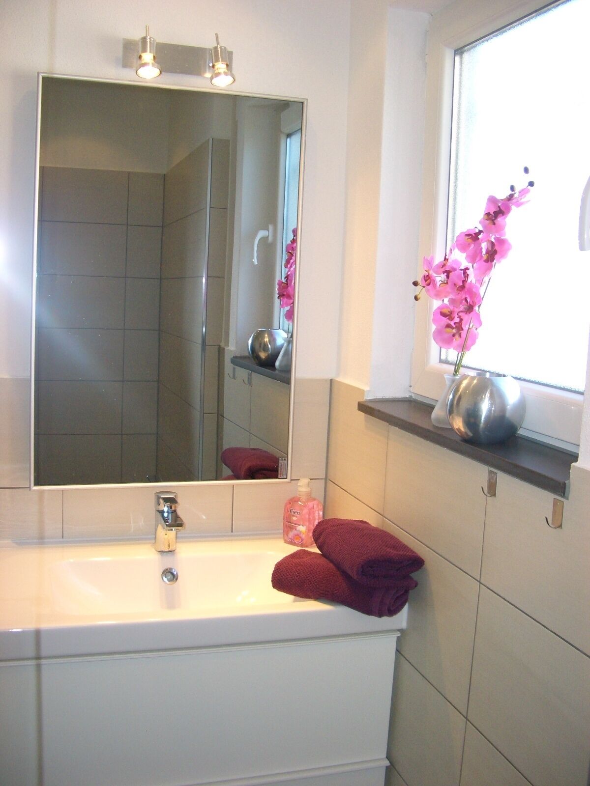 A bathroom with a large mirror, a sink, and a window with a vase of flowers.