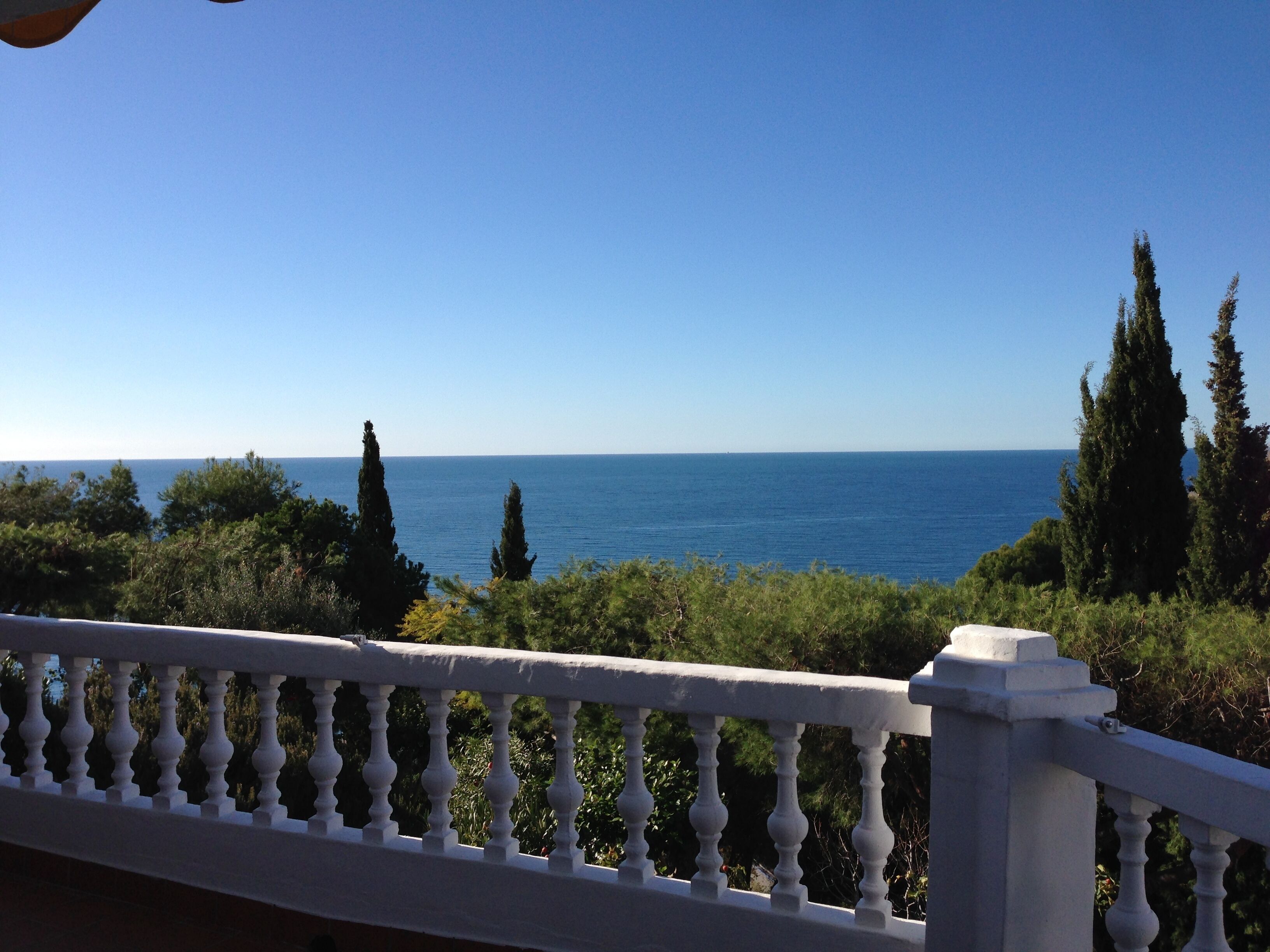 A balcony with a white balustrade overlooking a clear blue sea and greenery.