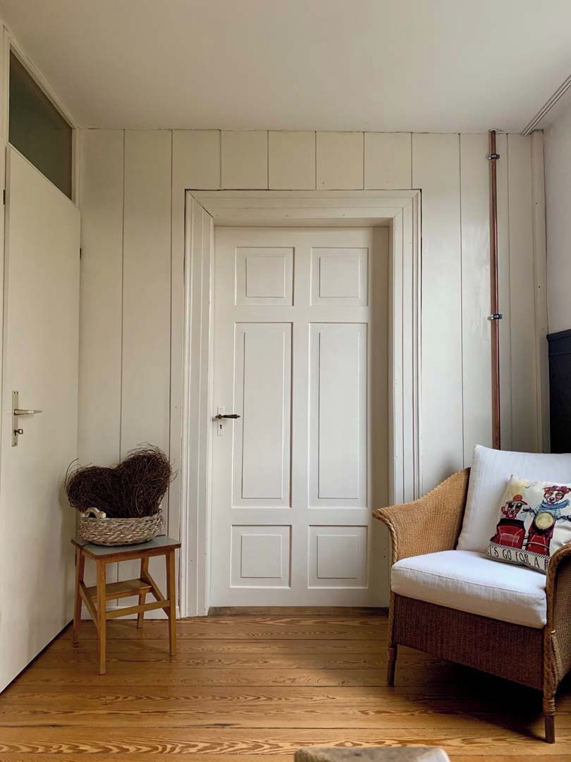 Una habitación con piso de madera, una puerta blanca, una cesta de mimbre sobre un taburete pequeño y una silla marrón con cojín.
